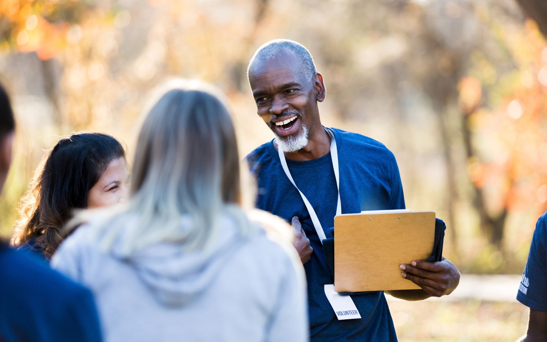 An older man with a gray beard and short hair, wearing a blue shirt and a volunteer badge, is smiling and talking to a group of people outdoors during daytime with autumn leaves in the background.